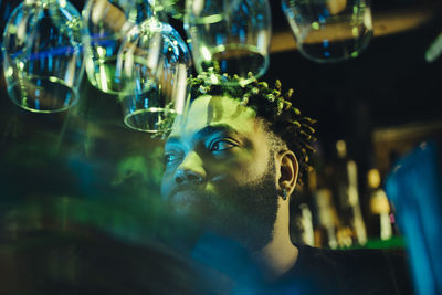 Young afro bartender looking away by wineglasses hanging upside down at illuminated bar