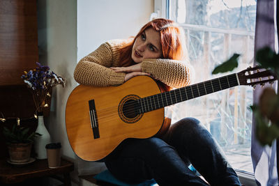 Unaltered candid portrait of young red haired woman in sweater playing acoustic guitar sitting by