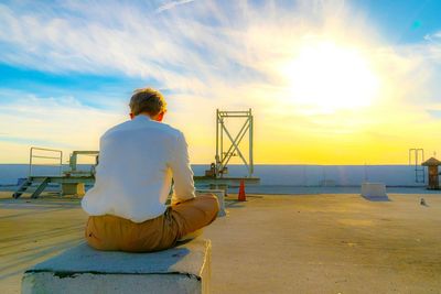 Rear view of man sitting on retaining wall against sky during sunset