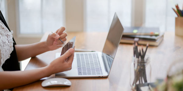 Midsection of woman using mobile phone over laptop on table