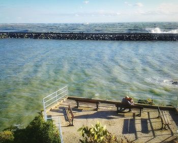 Rear view of man sitting on railing by sea against sky