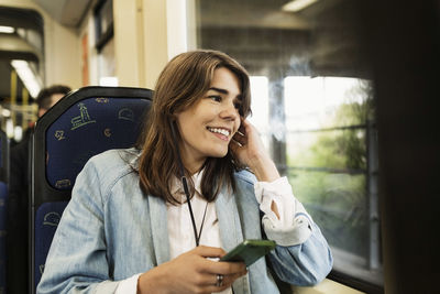 Smiling young woman holding mobile phone while looking through tram window