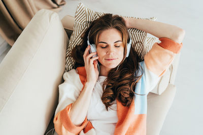 Young dreamy enjoying freckled redhead woman lying on sofa listening to music with headphones.