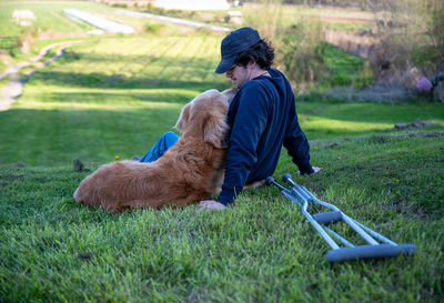 Big golden retriever dog cuddles in grass on rural hillside with young man with crutches on ground