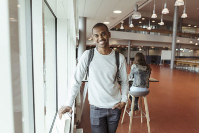 Portrait of smiling young student with backpack standing at university cafeteria with friend in background