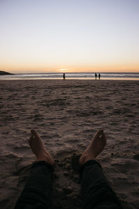 Low section of person relaxing on beach against sky during sunset