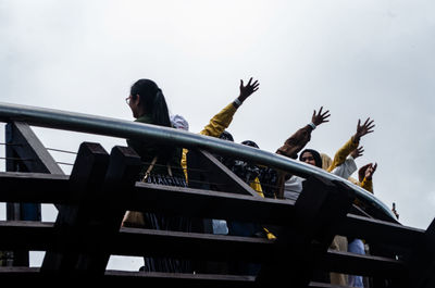 Low angle view of women with arms raised by railing against sky