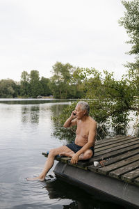 Senior man sitting on jetty at a lake talking on cell phone