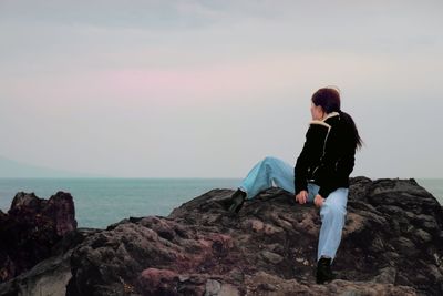 Woman looking at sea while sitting on rock against sky