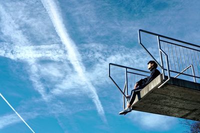 Low angle view of man sitting on built structure against blue sky