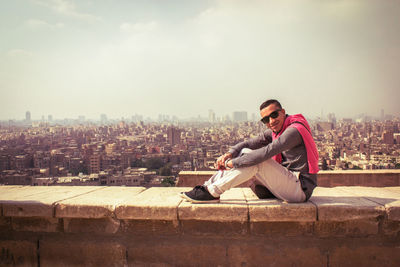 Portrait of man sitting on retaining wall against cityscape