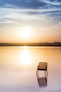 Chair on lake against sky during sunset