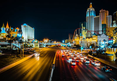 Light trails on city street by buildings against sky at night