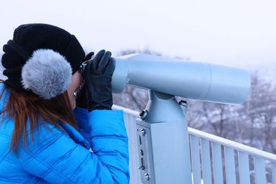 Side view of mid adult woman looking through coin-operated binoculars against sky during winter