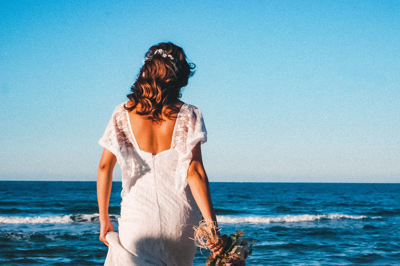 sea, water, sky, horizon over water, one person, horizon, blue, adult, beach, women, land, nature, clear sky, hairstyle, young adult, dress, female, leisure activity, standing, photo shoot, beauty in nature, three quarter length, vacation, long hair, trip, rear view, lifestyles, holiday, fashion, ocean, day, copy space, clothing, sunny, sunlight, scenics - nature, tranquility, outdoors, waist up, summer, casual clothing, wave, tranquil scene, person, travel, motion, relaxation, looking