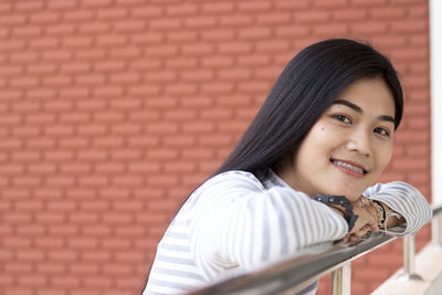 Portrait of young woman leaning on railing against brick wall