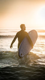 Man with surfboard wading in sea against sky during sunset