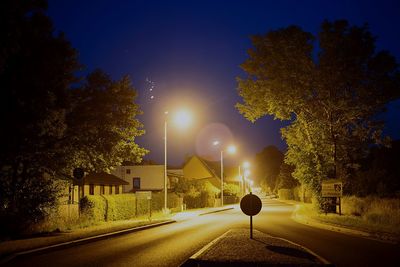 Illuminated street lights on road by houses against sky at dusk