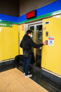 Back view full body of male with blindness in medical mask calling elevator standing with guide dog