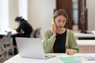 Middle-aged woman busy with documents talking on cellphone working in campus open space. education.