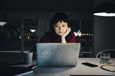 Dedicated young businesswoman with hand on chin using laptop while working late at office