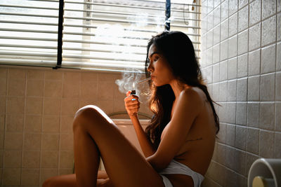 Side view of shirtless young woman lighting cigarette in bathroom