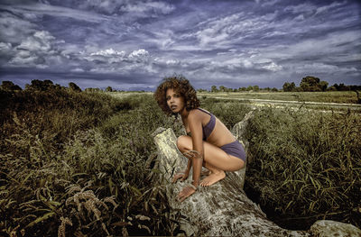 Portrait of woman crouching on fallen tree against cloudy sky