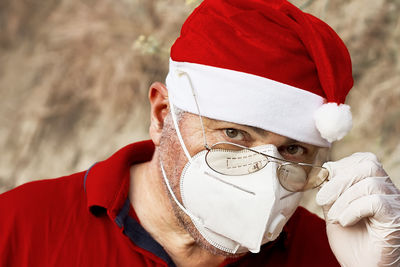 Mature man in santa claus hat, protective medical mask and gloves looks curious over his glasses. 