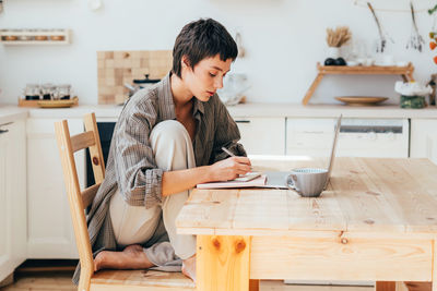 Young short-haired modern european woman works at home on a computer sitting at a wooden table.