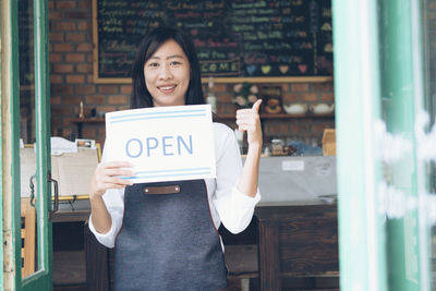 Portrait of female owner holding open sign while standing at doorway in cafe