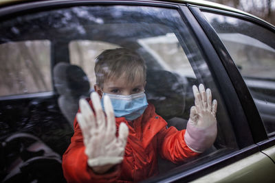 Little boy wearing respirator mask and medical gloves looking through a car window, stay safe