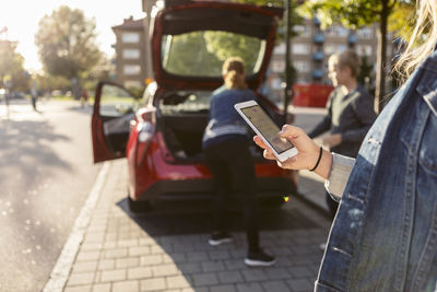 Midsection of teenager texting while mother and son standing near car trunk