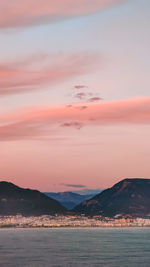 Pink clouds in evening sky over blue sea on background of city and mountains on horizon, vertical 