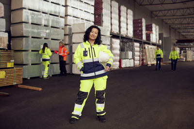 Portrait of smiling female worker in protective workwear holding hardhat while standing in factory