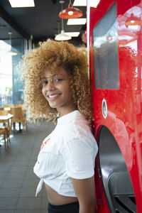 Young african american female with curly hair leaning on red soda machine in cafe and looking at camera