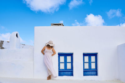 Unrecognizable female in summer clothes covering face with hat while standing outside rural white house with blue window shutters in sunny country