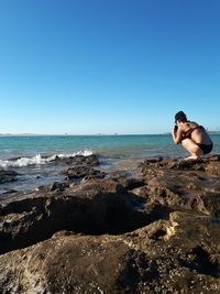 Woman in bikini photographing while crouching at seashore against clear sky