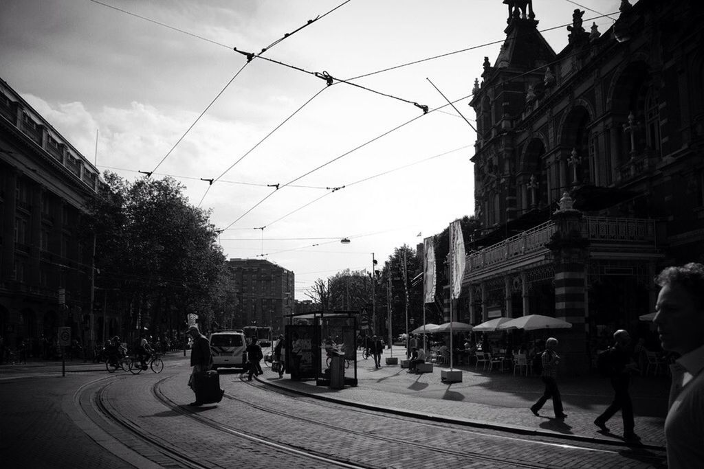 building exterior, architecture, built structure, power line, city, transportation, sky, street, city life, cable, incidental people, city street, railroad track, car, mode of transport, large group of people, road, men, electricity pylon