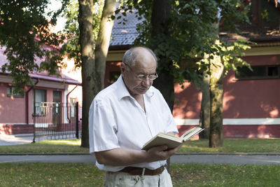 An elderly, bald man in a white shirt is reading a book in the park.
