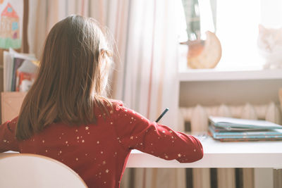 Pretty school girl with long hair in red dress draws with pencil at table in children's room at home