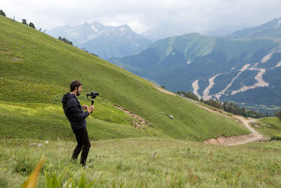 Guy kameraman in a jacket stands in the mountains on a green meadow and shoots a video on steadicam