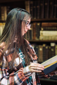 Student with glasses holding a large old book and reading against the background of bookshelves 