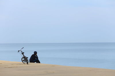 Woman with bicycle sitting at beach against clear sky