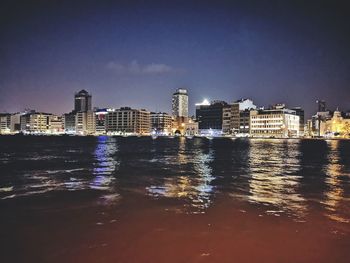 Illuminated buildings by sea against sky