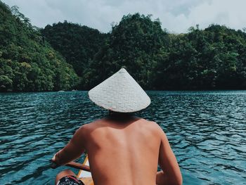 Rear view of shirtless man in boat on lake against trees