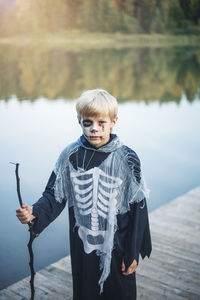Portrait of smiling boy standing against lake in halloween costume with painting face 