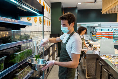 Side view of male seller in apron and mask putting green herbs in bowl while working in salad bar in supermarket