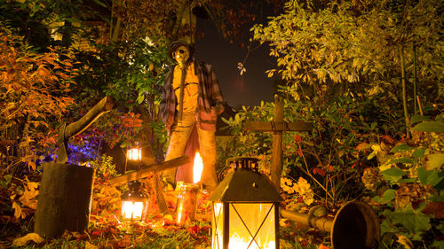 Scarecrow with gas mask amidst trees and illuminated lanterns at night