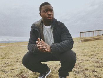 Full length of man looking away while crouching on grassy field against sky