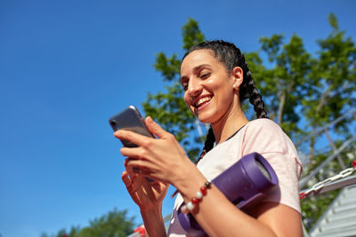 Smiling young woman using smart phone against blue sky